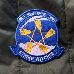 Könnte beinhalten: Ein blauer und weißer gestickter Aufnäher mit dem Text "501st Joint Fighter Wing Strike Witches". Der Aufnäher zeigt einen fünfzackigen Stern, über dem sich vier gelbe Besen kreuzen.
