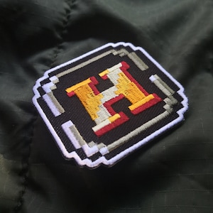 Puede incluir: Un parche bordado pixelado con un borde blanco, que presenta una gran letra amarilla "H" con contornos rojos sobre un fondo negro.