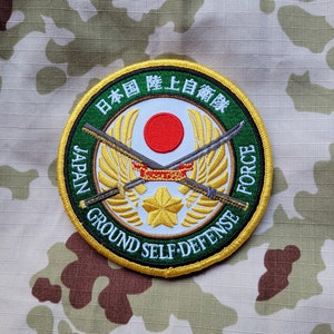 Pode incluir: Um remendo redondo branco com uma borda verde e uma guarnição dourada. O remendo apresenta um círculo vermelho com uma estrela branca no centro, duas espadas cruzadas e o texto "JAPAN GROUND SELF-DEFENSE FORCE" em um círculo dourado ao redor do exterior. O texto "日本国陸上自衛隊" está escrito em caracteres japoneses ao redor da parte superior do remendo.