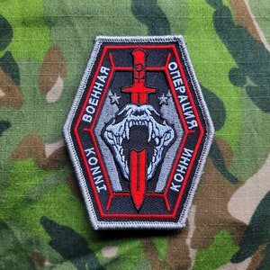 Könnte beinhalten: Ein grauer, sechseckiger Patch mit roten Nähten. Der Patch zeigt ein schwarzes und rotes Schwert mit einem weißen Schädel und Zähnen. Der Text "ВОЕННАЯ ОПЕРАЦИЯ" ist in Schwarz um den Rand des Patches geschrieben.