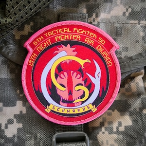 May include: A pink embroidered patch with a black and white skull and snake design. The text on the patch reads "67th Tactical Fighter Sq 13th Night Fighter Air Division Schwarze".