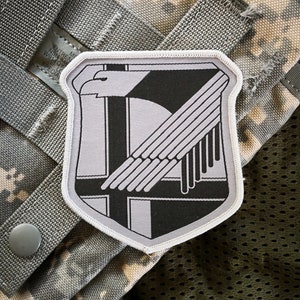 May include: A white and gray embroidered patch with a stylized eagle design. The eagle is facing right and has a wing spread. The patch has a black border and a white outline.
