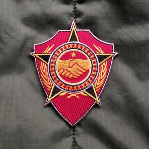 May include: An embroidered patch in red, white and gold, featuring a star, a handshake and a wreath. The patch is shield-shaped with a black outline.