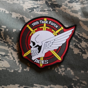 Könnte beinhalten: Ein gestickter schwarzer und roter Aufnäher mit einem Totenkopf- und Knochenkreuz-Design. Der Aufnäher trägt den Text "19th Task Force Aces".