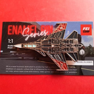 May include: A silver and red enamel pin of a fighter jet with the text "ENA Series" and "1:1 Scale Product" on the card. The card also has the text "FEI CORP" and "MODEL/NUMBER".