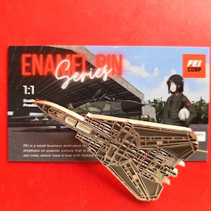 May include: A gold-colored enamel pin of a fighter jet with a detailed design. The pin is on a card with the text "ENAMEL PIN Series" and "FEI CORP".