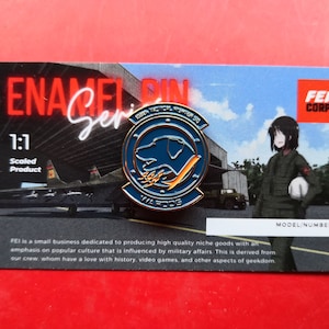 May include: A blue and gold enamel pin with a dog head and the text "189th Tactical Fighter Sq" and "WAR DOGS". The pin is on a white card with the text "ENAMEL PIN Series" and "FEI CORP".