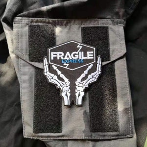 Könnte beinhalten: Ein schwarz-weißes Patch mit einem weißen Skelett-Hand-Design, das ein Hexagon mit dem Text "FRAGILE EXPRESS" in weißen Buchstaben mit einem blauen Blitz hält.