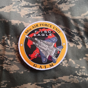 Może przedstawiać: Naszywka haftowana na żółto i pomarańczowo z białym obramowaniem. Naszywka przedstawia szary myśliwiec F-15C Eagle z czerwono-biało-niebieskim ogonem. Tekst "66th AIR FORCE UNIT" znajduje się nad samolotem, a "F-15C EAGLE" pod samolotem. Tekst "GAIM" znajduje się pod samolotem.