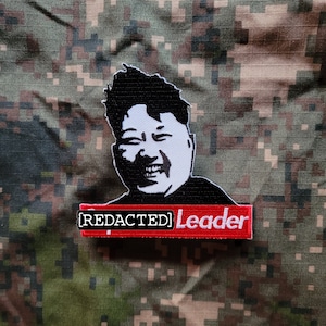 May include: A red and white embroidered patch with a black and white image of a man with a large smile. The patch says "[REDACTED] Leader" in white text on a red background.