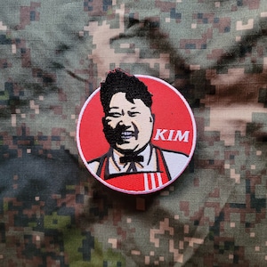May include: A red and white embroidered patch with a cartoon image of a man with black hair and a mustache. The man is wearing a red apron and the text "KIM" is written in white on the patch.