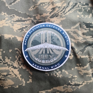 May include: A blue and white embroidered patch with a space shuttle and the text "Space Elevator Defense System", "Liberty", "ISEV Justice", and "Arsenal Bird".