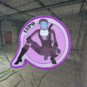 Op de afbeelding: Geborduurde patch met een zittende futuristische figuur, met de naam "LUPO" erboven. De patch heeft een paarse rand en een lichtpaarse achtergrond. De figuur draagt een masker en donkere kleding.