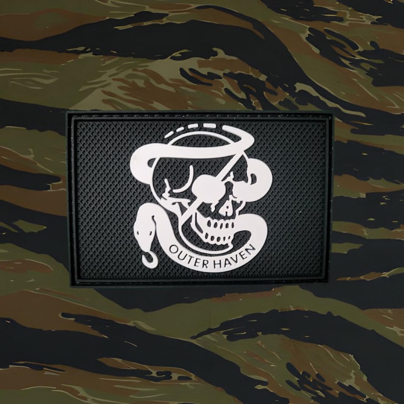 Op de afbeelding: Zwart-witte patch met een schedel- en slangontwerp. De tekst "Outer Haven" staat onder de schedel. De patch is op een camouflage achtergrond.