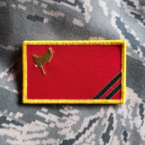 May include: A red embroidered patch with a yellow border and black stripes. A gold bird is in the upper left corner.