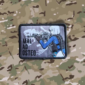Könnte beinhalten: Ein gestickter Patch in Schwarzweiß mit einer Frau, die ein Gewehr hält. Die Frau trägt einen blauen Overall und einen weißen Helm. Der Text "MAL AD OSTEO" ist auf dem Patch sichtbar.