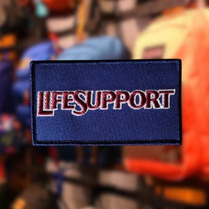 May include: A rectangular navy blue patch with the embroidered text "LIFESUPPORT" in white with red accents. The patch has a black border and is likely designed to be sewn onto clothing or gear.