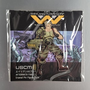 Pode incluir: Um broche de esmalte verde e amarelo com um personagem da franquia Alien. O personagem é um soldado que usa um capacete e segura uma arma. O broche é etiquetado com "USCM" e "エイリアンVSプレデ" com o texto "AFTERMATH FAN CLUB" e "Enamel Pin Figure Type".