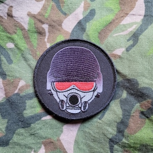 Op de afbeelding: Zwart-witte geborduurde patch van een persoon met een gasmasker met rode ogen. De patch is rond en heeft een zwarte rand.