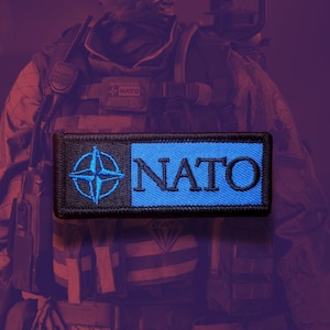May include: A rectangular embroidered patch featuring a black section on the left with a blue four-pointed compass star emblem and a blue section on the right with the word NATO embroidered in black capital letters with a black border.