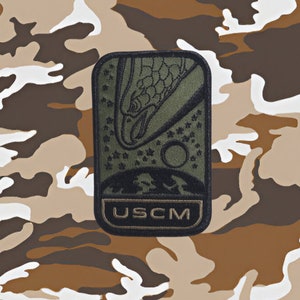 May include: A green and black embroidered patch with a stylized eagle design and the text "USCM".