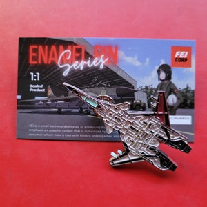 May include: A silver and black enamel pin of a fighter jet with a red tail. The pin is 1:1 scaled and features the text "ENA Series" and "FEI CORP".
