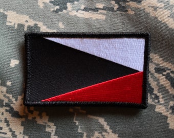Puede incluir: Parche negro, blanco y rojo con un diseño diagonal. El parche está cosido sobre una tela de camuflaje.