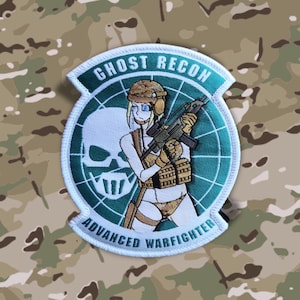 Könnte beinhalten: Ein weißer gestickter Aufnäher mit einem grünen Kreis in der Mitte. Der Kreis zeigt einen Totenkopf mit Knochen, eine Frau, die ein Gewehr hält, und den Text "Ghost Recon Advanced Warfighter".