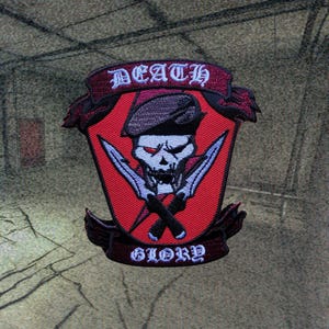 Puede incluir: Parche bordado rojo y blanco con una calavera con boina y dos cuchillos. El parche tiene las palabras "DEATH" y "GLORY".