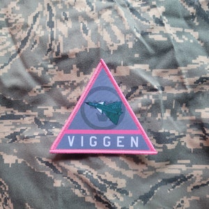 Peut inclure: Un écusson triangulaire rose avec un fond gris. L'écusson représente un avion de chasse stylisé et le mot "VIGGEN" en lettres blanches.