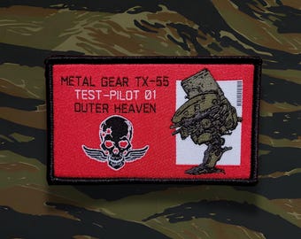 May include: A rectangular patch with a red background featuring the text "METAL GEAR TX-55 TEST-PILOT 01 OUTER HEAVEN." It also displays a skull with wings and a detailed illustration of a military vehicle. The patch is bordered in black.