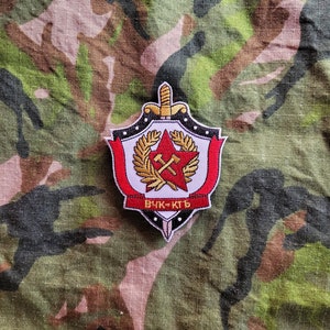 Peut inclure: Un écusson brodé rouge et blanc avec une étoile rouge, des marteaux et une pioche croisés et une épée. L'écusson a une bordure noire avec des points blancs et le texte "ВЧК-КГБ" en rouge.