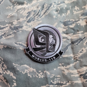 Könnte beinhalten: Ein gestickter Aufnäher in Schwarz und Silber mit einem stilisierten "M" und dem Text "MOBIUS-118".