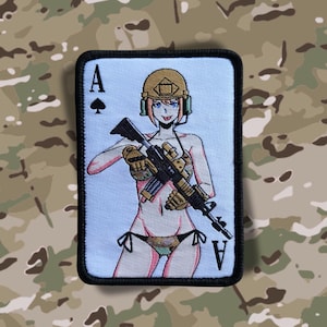 Puede incluir: Un parche bordado azul y blanco con una imagen de dibujos animados de una mujer en bikini sosteniendo un rifle. El parche tiene la letra 'A' y un símbolo de pica en la esquina superior izquierda y la letra 'A' en la esquina inferior derecha.