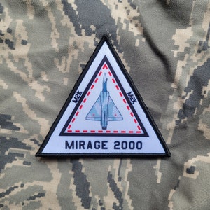 May include: A white triangular patch with a black border. The patch features a silhouette of a Mirage 2000 fighter jet in flight. The text "MIRAGE 2000" is printed in black below the jet. The text "MK" is printed in black on each side of the jet. A red dashed line surrounds the jet.