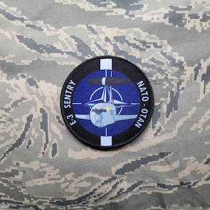 May include: A black and blue circular patch with a white border. The patch features a white airplane with a red nose and a blue radar dish. The text "E-3 SENTRY NATO - OTAN" is printed around the airplane.