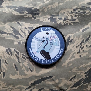 May include: A round, black and white patch with the text "AIRFORCES LITTLE HELPER" and "E-737" around a cartoon illustration of a person wearing a blue jacket and black trousers, holding a pair of glasses and standing in front of a plane.