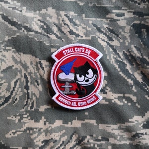 Könnte beinhalten: Gestickter Aufnäher mit rotem Rand und dem Text "STALL CATS SQ" oben. Der Aufnäher zeigt eine Zeichentrickkatze, eine blau-weiße Rakete und den Text "9000th AD, 69th WING". Der Aufnäher befindet sich auf einem Tarnhintergrund.