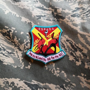 May include: A colorful embroidered patch with a red and yellow shield and a sword. The shield has a red and yellow cross with a red devil holding a spear. The text "ESPADA" is above the shield. The text "9th Air & Land Div 11th Tac Ftr SQ" is below the shield.