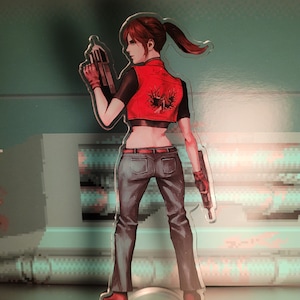 May include: A red and black anime character figurine with a red vest and blue jeans. The character is holding a gun in their right hand and has a ponytail. The figurine is standing on a clear plastic base.