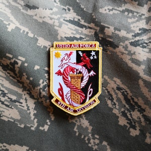 以下が含まれることがあります： 上部に「USTIO AIR FORCE」の文字が入った軍用パッチ。パッチは白い縁取り、黄色と黒の背景、赤いドラゴンが特徴のシールドデザインです。「6TH AIR DIVISION」の文字が下部にあります。