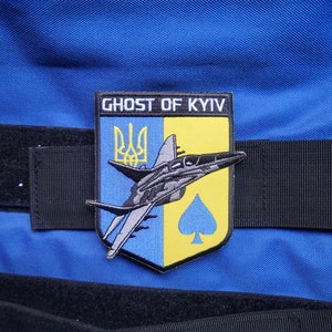 Pode incluir: Um remendo azul e amarelo com o texto "Ghost of Kyiv" e uma silhueta preta de um caça. O remendo também apresenta o tridente ucraniano e um paus preto.