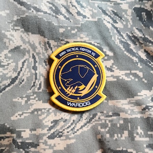 May include: A yellow and blue embroidered patch with the text "108th Tactical Fighter SQ" and "WARDOG". A black silhouette of a dog is in the center of the patch.