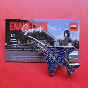 Könnte beinhalten: Eine blaue und silberne Emaille-Anstecknadel mit einem Kampfjet. Die Anstecknadel ist ein 1:1-Modell und Teil der "Enamel Pin Series".