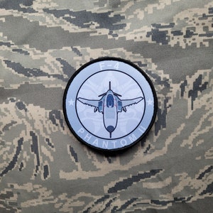 May include: A blue and white embroidered patch with a gray F-4 Phantom jet in the center. The patch has a black border and the text "F-4 PHANTOM II" around the edge.
