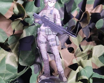May include: A clear acrylic standee featuring an anime-style soldier in a camouflage uniform, holding a rifle. The figure has blonde hair and is wearing a helmet and goggles. The standee is placed in front of a camouflage net background.