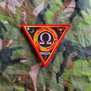 Op de afbeelding: Een driehoekige patch met een rode rand en een zwarte achtergrond. De patch heeft een gouden Omega-symbool, het woord "OMEGA" in het wit en een bliksemschicht. De patch heeft ook een maan, sterren en een rood-geel ontwerp.