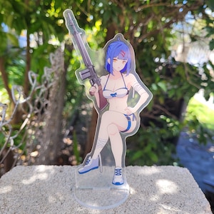 Könnte beinhalten: Ein durchsichtiger Acryl-Ständer mit einer Anime-Figur mit blauen Haaren, die ein Gewehr hält. Die Figur trägt einen blauen und weißen Bikini und hat einen Verband am Arm. Der Ständer steht auf einer grauen Betonfläche.