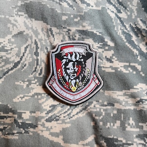 Peut inclure: Un écusson brodé gris, noir et rouge avec une tête de lion et le texte "Scarface 371st FTW 1st Squadron".