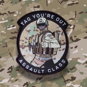 May include: A circular patch featuring a soldier in combat gear, with the text "TAG YOU'RE OUT ASSAULT CLASS" in a black border. The soldier is depicted in tan and brown tones, set against a camouflage background.
